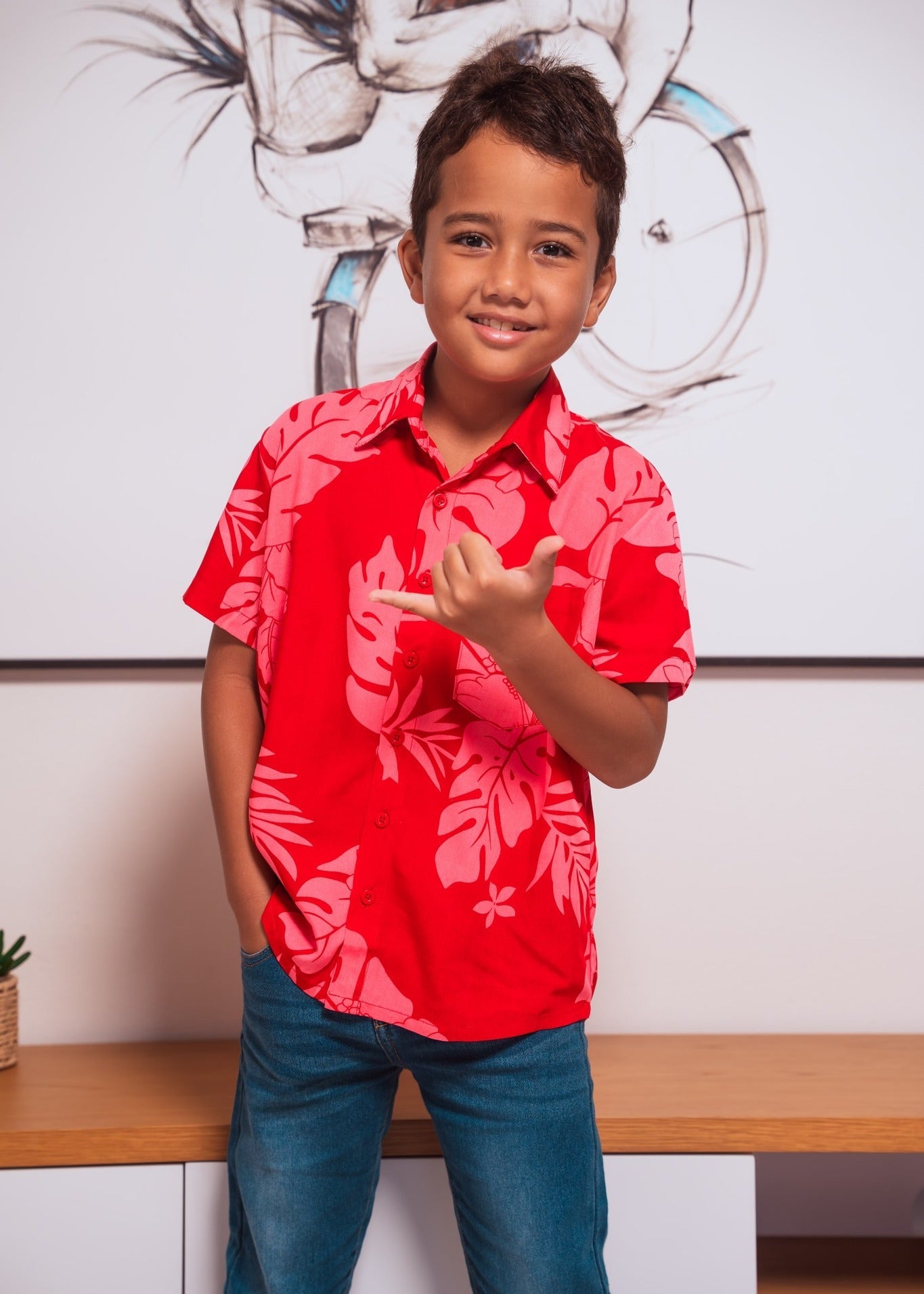 Young boy from tahiti wearing a red Polynesian floral Bamboo Kultur shirt, smiling and making a Hang loose sign