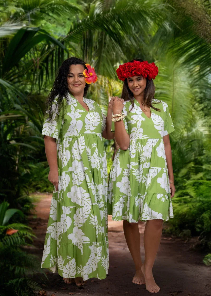 Two women wearing Bamboo Kultur’s “Vaihere” green floral Polynesian dress, one in the long version and the other in the short version, standing in a tropical setting.