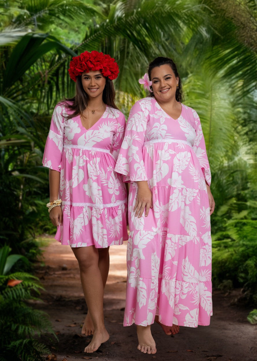 Two women wearing Bamboo Kultur’s “Merahi” pink floral Polynesian dress, one in the long version and the other in the short version, standing in a tropical setting